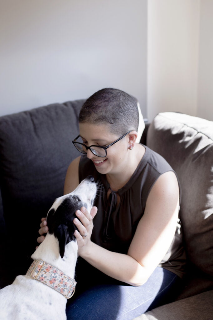 Person with short hair and glasses smiles and pats a black and white greyhound